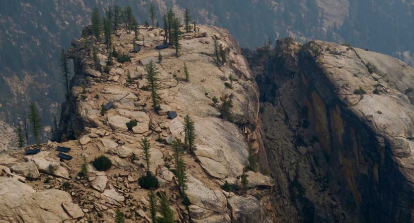 From an aerial point of view, a couple of tents rest on a cliff high above the landscape below. A few evergreen trees also dot the cliff. 